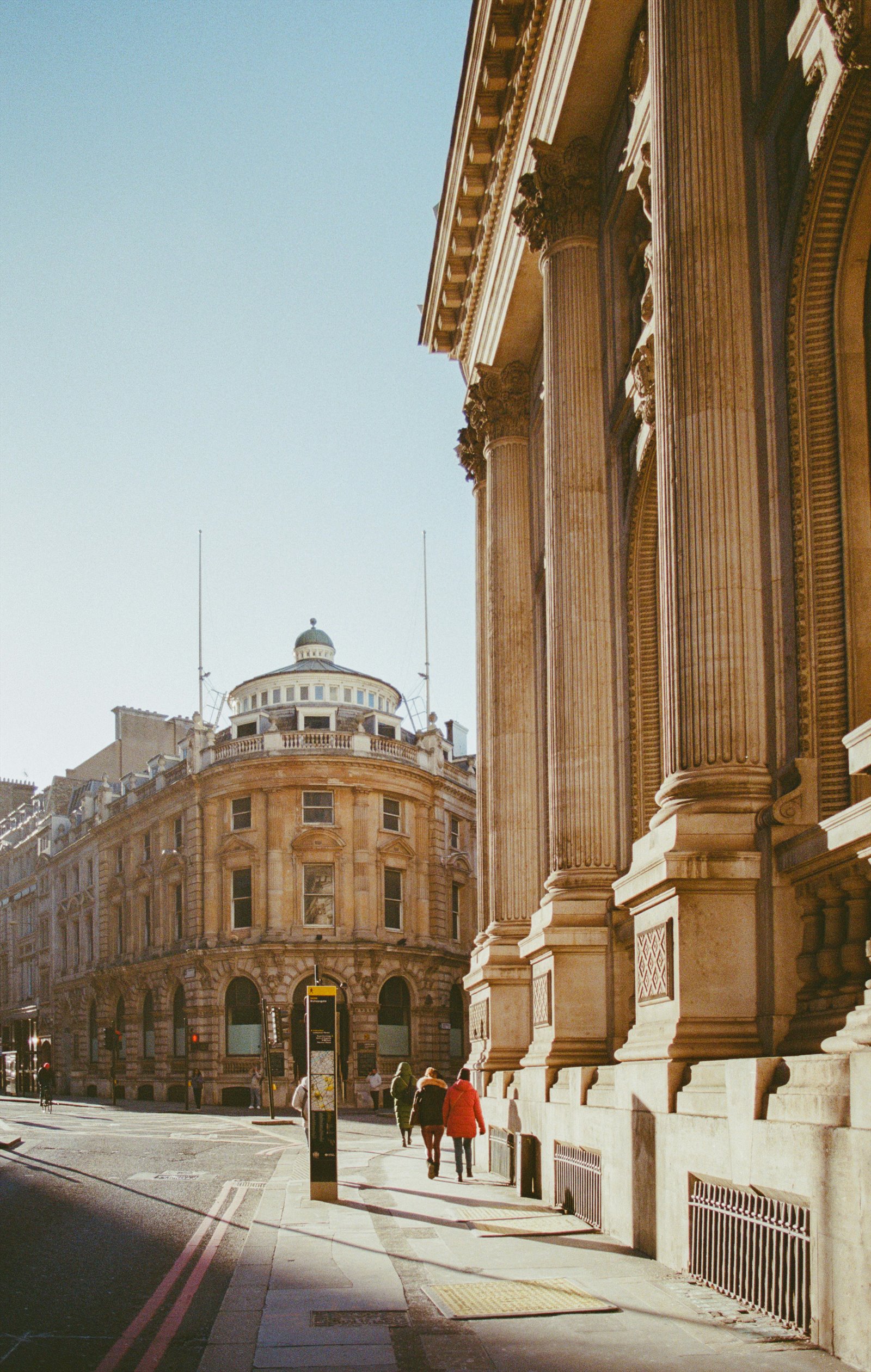 People walking along a sunlit City of London street beside classical stone façades.